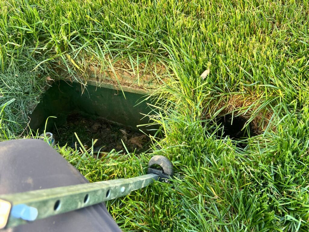 A large metal box holding in-ground sprinkler equipment is buried in a lush green lawn.